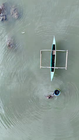 Aerial drone view of a man swimming near a traditional boat with a floating basket in shallow water in the Philippines. Vertical
