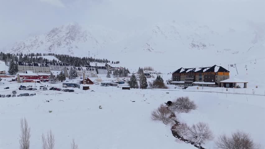 Breathtaking aerial establishing shot of Las Leñas ski resort in Mendoza, Argentina, featuring snow-covered hotels and majestic Andean mountain peaks on a cold winter day.