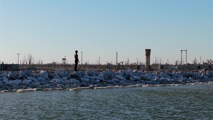 A person strolls along a rocky embankment in Villa Epecuén, Buenos Aires, Argentina, beside a large body of water.