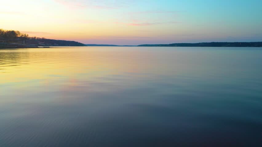 Sunset aerial over Smith Mountain Lake, Virginia, with calm water, wooded shoreline, distant lake homes, pastel sky, and gentle golden reflections across the wide reservoir.