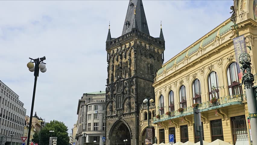 Street view of the Gothic Powder Tower in Prague, Czech Republic, beside the ornate Municipal House, with historic architecture, tall black spire, city street signs, lampposts, and cloudy sky.