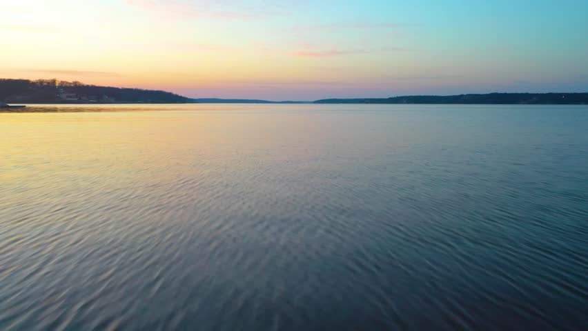 Wide drone view above Norris Lake, Tennessee at dusk, featuring calm rippled water, forested shoreline, distant coves, soft sunset light, and peaceful reservoir scenery in evening haze.