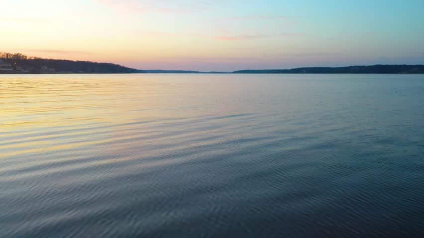 Forward boat POV on a quiet lake at dusk, showing gentle surface ripples, distant tree-lined shoreline, soft sunset colors, and calm open water glowing beneath a pastel evening horizon.