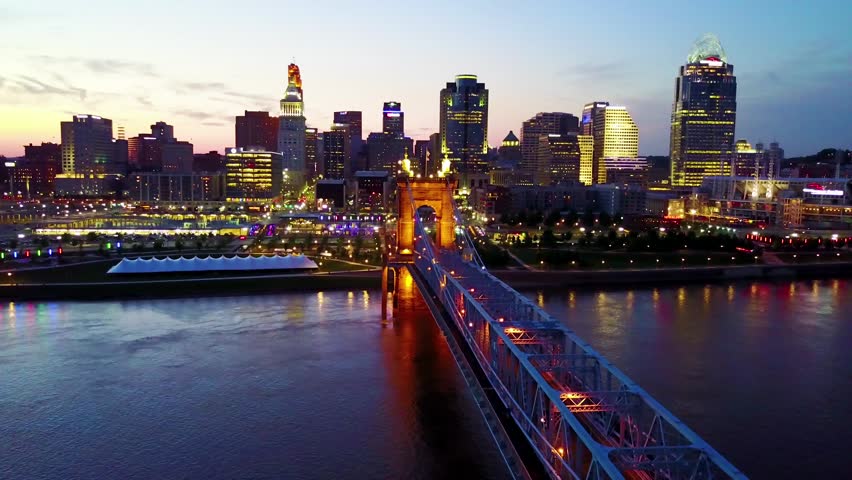 Aerial view showing Cincinnati skyline at twilight. Cinematic drone footage captures Roebling suspension bridge and illuminated skyscrapers reflected in Ohio river during blue hour transition.