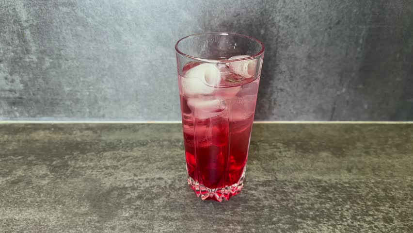 A clear glass filled with a vibrant red beverage and numerous ice cubes sits on a dark grey stone-textured countertop against a grey textured background. A metal stir stick is placed inside the drink