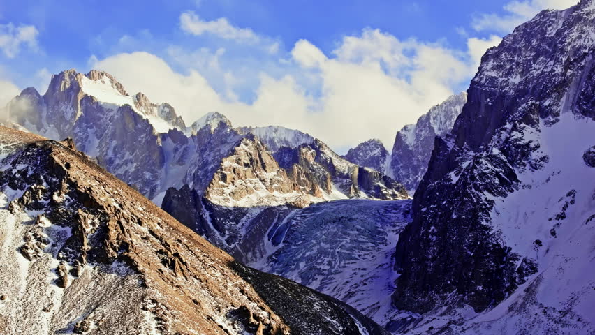 High-altitude mountain wilderness features majestic snow-capped peaks and a glacier. Clear blue sky enhances the serene and awe-inspiring atmosphere. Ascending drone view.