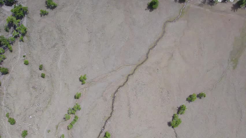 Aerial riverbank view showing reflective river surface beside dry pebble floodplain, shoreline structures, scattered vegetation, and winding drainage channel beneath bright daylight