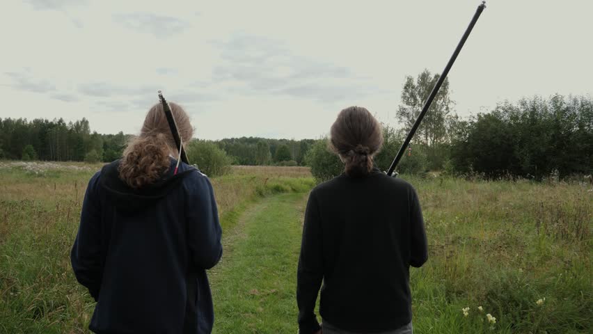 Rear view of two people walking along a narrow grassy path through a wildflower-strewn meadow toward distant mixed trees under an overcast sky. Both carry fishing rods