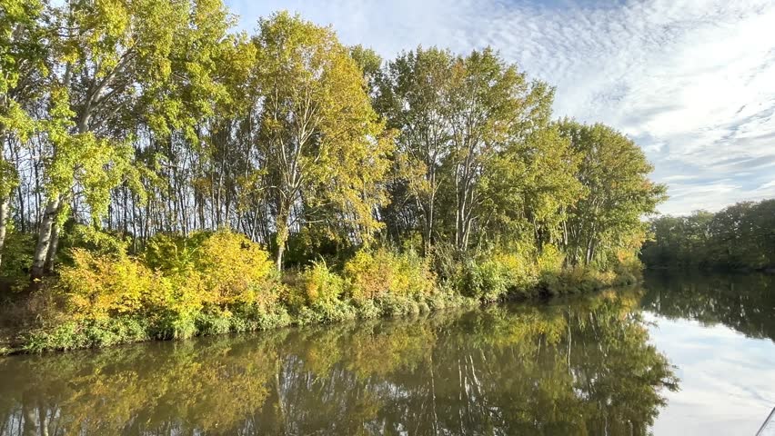 calm river, yellow-green leaves on the trees, an idyllic river landscape seen from a boat in autumn, sunny day, blue sky, church in the background, trees reflected in the river, green reflection