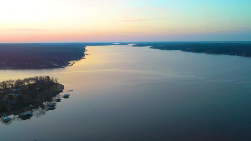 Drone footage over Beaver Lake, Arkansas at sunset, showing calm blue water, wooded Ozark shoreline, distant homes, pastel evening sky, and warm reflections across the reservoir.
