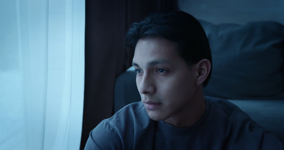 Alone man sit floor by window at night. Sad young person looking out in dark room at home. Gloomy atmosphere, loneliness, depression, solitude. Melancholy mood, introspection, psychological struggle.