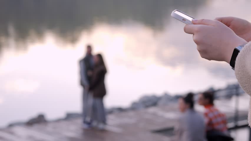 Close-up of hands holding a phone, capturing a blurred scene by water. Background shows people relaxing near a lake at sunset. Calm, peaceful mood with warm golden light. Ideal for social media