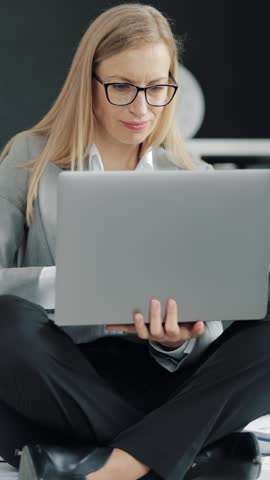 A woman sits on the floor in an office and works on a laptop with focus and interest.