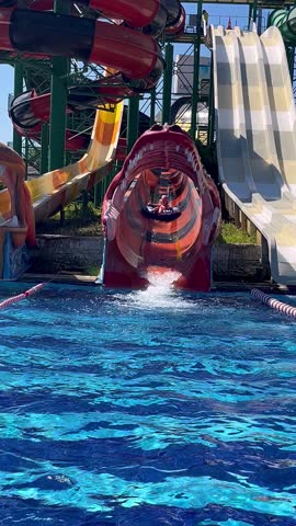 A red water slide tunnel ending in a blue pool at a water park with child splashing into the water, summer fun, waterpark excitement, outdoor recreation, joyful splash, vacation thrill.