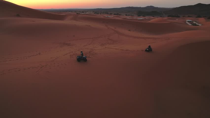 Quad bikes traverse the Erg Chebbi dunes near Merzouga, Errachidia, Drâa-Tafilalet, Morocco, Africa, leaving tracks across the Sahara during warm sunset light, drone pan tracking shot.