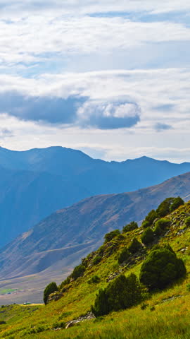 Time lapse of clouds over mountain valley in Kyrgyzstan. Foreground green hillside with shrubs contrasts with distant ridges and soft light on slopes.