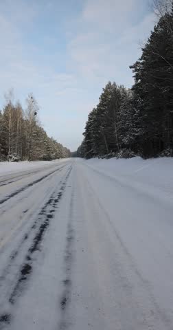paved road covered with ice for cars through a winter forest in the snow , a track in the forest in the winter season, different types of trees grow on the side of the road against the blue sky