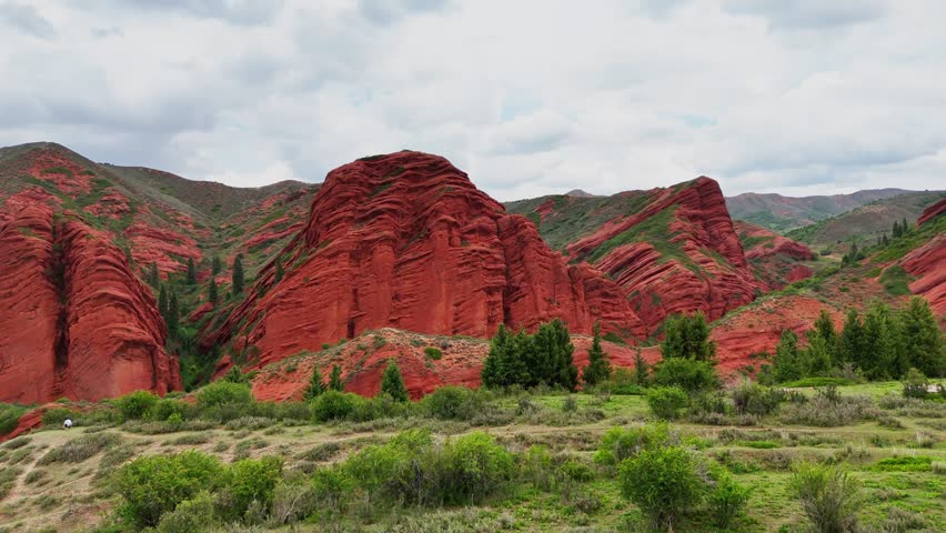Drone left‑to‑right lateral pass along vivid red layered sandstone cliffs with green hills, overcast