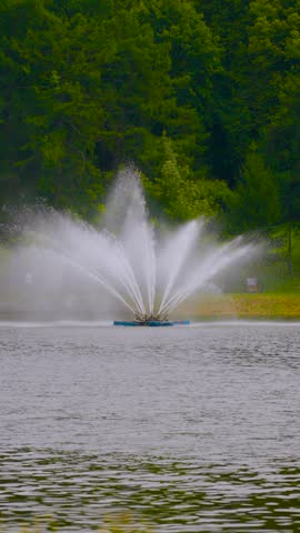 Fountain Water Spray In Lake Surrounded By Green Forest In A Tranquil Park Setting.