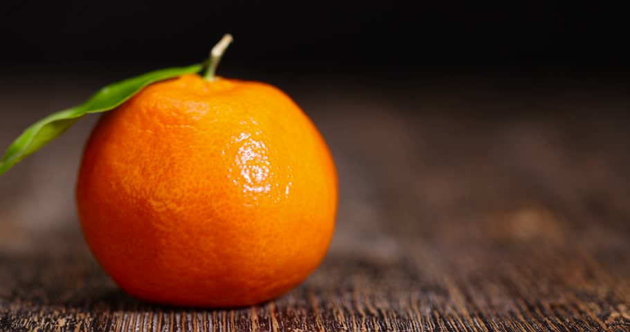 juicy orange tangerine with one green leaf, fruit on a wooden table, unpeeled ripe orange-skinned small tangerine with a long green leaf