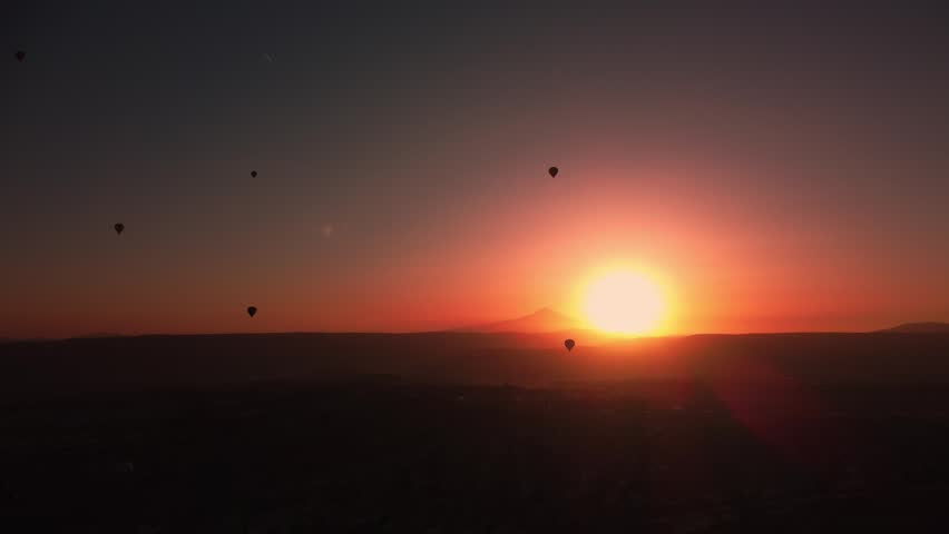 Aerial view of hot air balloons illuminated by sunset glow over rugged landscape. Drone footage of aerostats flight over mountain ridge. Evening scene with dusk above rocky landscape. Travel theme