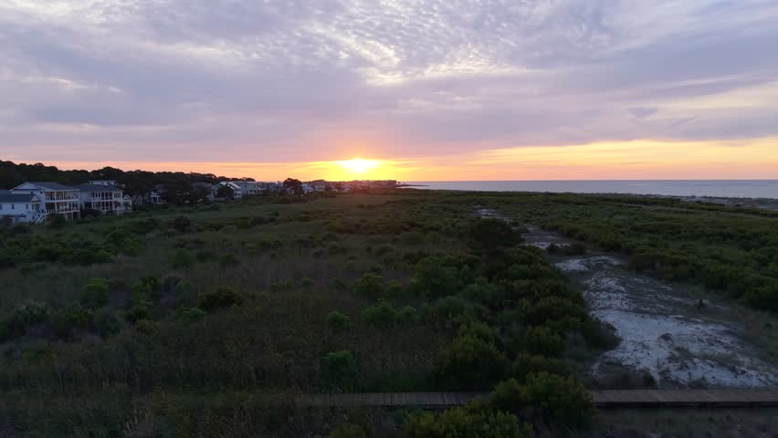 Low-altitude aerial view of a wooden beach access boardwalk winding through coastal sand dunes and maritime shrubs during a vibrant Atlantic sunrise on Fripp Island, South Carolina.