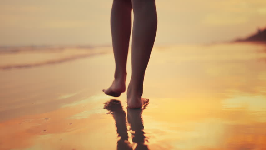 Close up of bare feet of young Asian woman walking on wet sand beach at sunset, lower body part of female strolling on sea shore during golden hour, tropical summer vacation, travel lifestyle.