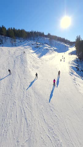 Smooth FPV drone flight over a snowy ski slope with skiers enjoying winter sports in the mountains on a clear sunny day.