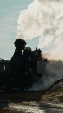 Vertical Screen: The Mocanita steam train stands at the station in Bucovina, Romania. Smoke billows from the locomotive as it prepares for an upcoming journey through the mountains.
