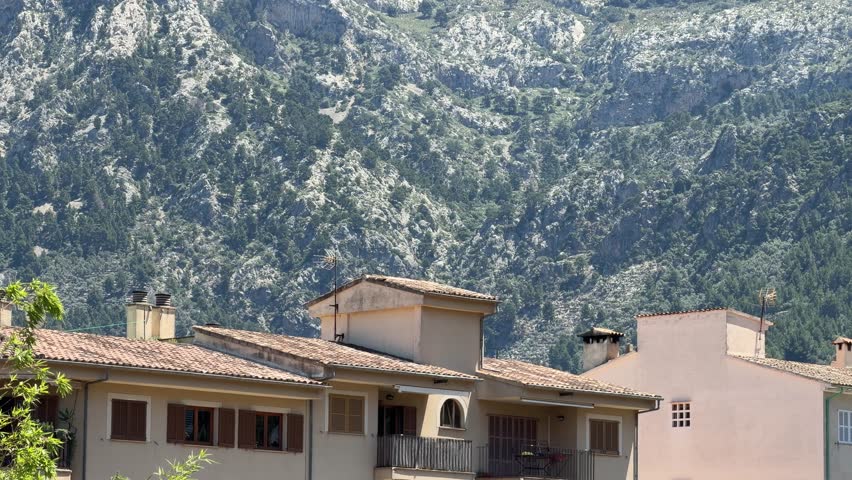 Traditional houses in Soller valley, Mallorca with terracotta roofs and shutters, framed by dramatic Tramuntana mountains under clear blue sky on a sunny day.