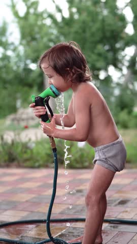 A young boy plays with a garden hose, spraying water. The boy smiles as he interacts with the hose in a lush green garden setting.
