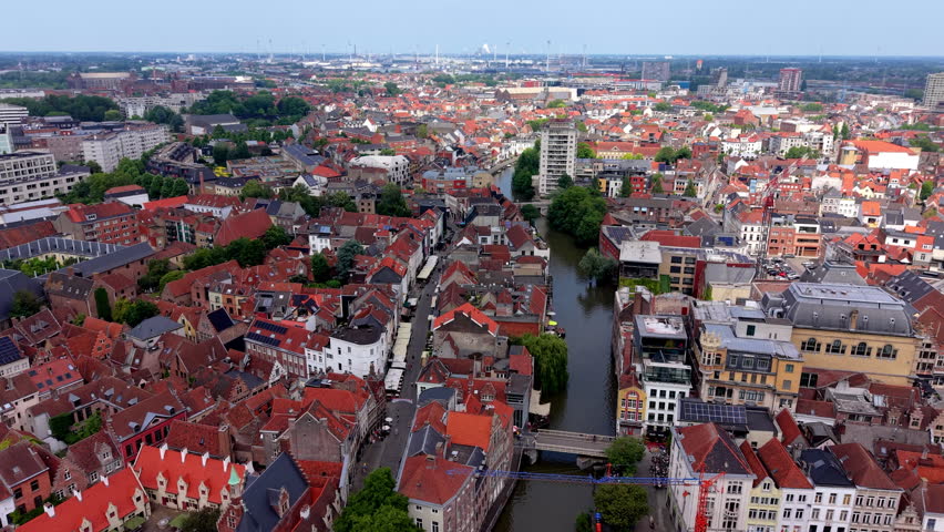 A comprehensive high-angle aerial drone photograph capturing the dense and vibrant cityscape of a historic town center in Western Europe. The frame is filled with an intricate tapestry of traditional buildings featuring steep red-tiled roofs, mixed with historical brick facades and modern residential complexes. A narrow, dark river canal cuts vertically through the center of the town, spanned by small pedestrian footbridges and bordered by lush green trees and waterfront buildings. An active pedestrian street runs parallel to the canal on the left, lined with outdoor umbrellas and shops. In the background, the expansive urban landscape stretches toward the horizon, revealing modern tower blocks, distant industrial wind turbines, and industrial infrastructure under a bright, clear daytime sky.