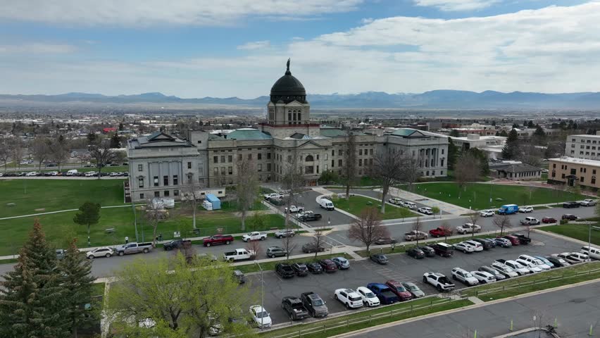 Montana State Capitol with copper dome rises above green lawns and full parking lots in Helena, with low-rise cityscape and Rocky Mountain foothills beyond. Montana, USA. Aerial drone approaching shot