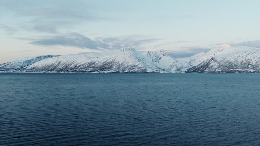 Snow-covered Arctic mountains rising beyond deep blue fjord waters, drone