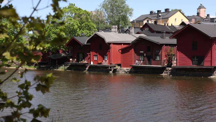 iconic red wooden riverside storehouses of Porvoo, Finland, framed from under a bridge. A small boat sits on the calm water, while the historic cathedral and old town buildings peak over the roofs