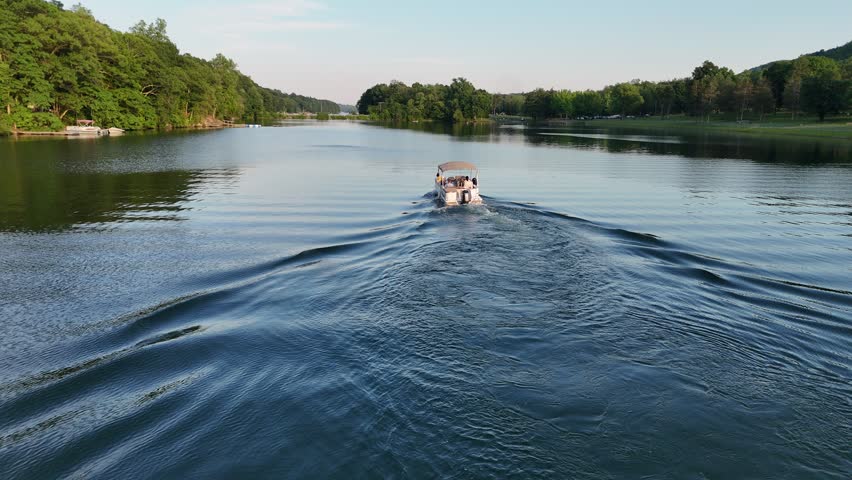 Stunning high-angle aerial drone footage of Squantz Pond State Park, located near Sherman and New Milford, Connecticut. The video clips showcase recreational pontoon boats cruising on the calm water surface during a warm golden hour sunset. The scenic New England landscape features a sandy public swimming beach, lush green forested hills, and brilliant sunlight reflecting off the lake. Perfect for summer vacation promos, regional tourism marketing, lifestyle lakeside living documentaries, and nature travel stock media.