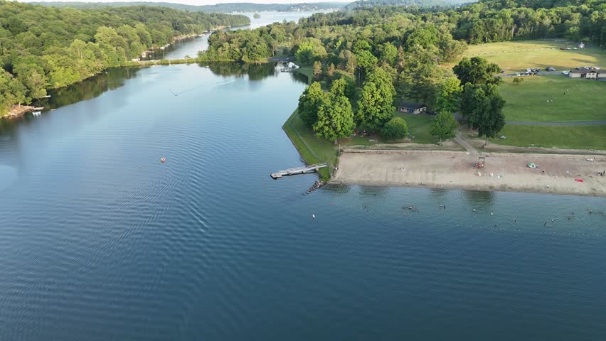 Stunning high-angle aerial drone footage of Squantz Pond State Park, located near Sherman and New Milford, Connecticut. The video clips showcase recreational pontoon boats cruising on the calm water surface during a warm golden hour sunset. The scenic New England landscape features a sandy public swimming beach, lush green forested hills, and brilliant sunlight reflecting off the lake. Perfect for summer vacation promos, regional tourism marketing, lifestyle lakeside living documentaries, and nature travel stock media.