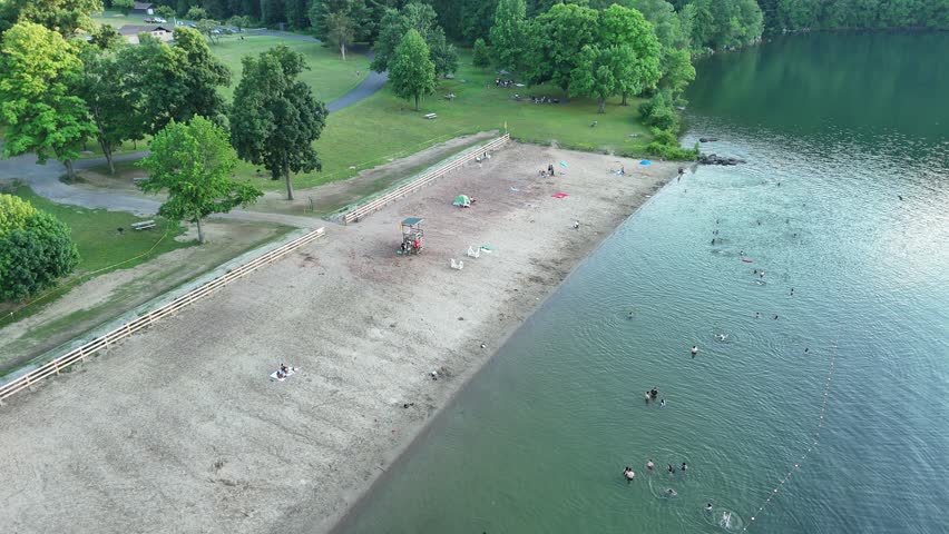 Stunning high-angle aerial drone footage of Squantz Pond State Park, located near Sherman and New Milford, Connecticut. The video clips showcase recreational pontoon boats cruising on the calm water surface during a warm golden hour sunset. The scenic New England landscape features a sandy public swimming beach, lush green forested hills, and brilliant sunlight reflecting off the lake. Perfect for summer vacation promos, regional tourism marketing, lifestyle lakeside living documentaries, and nature travel stock media.