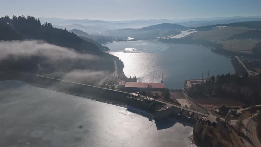 Niedzica-Zamek hydroelectric dam on Lake Czorsztynskie in Lesser Poland, with frozen reservoirs, forested hills and mountain valley landscape in aerial view.