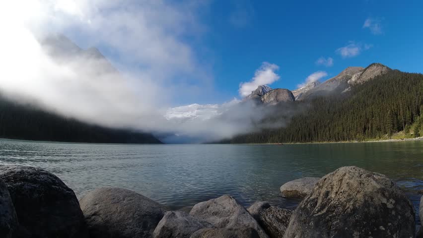 Low clouds drift away to reveal a turquoise alpine lake framed by forested peaks and distant glaciers in timelapse.