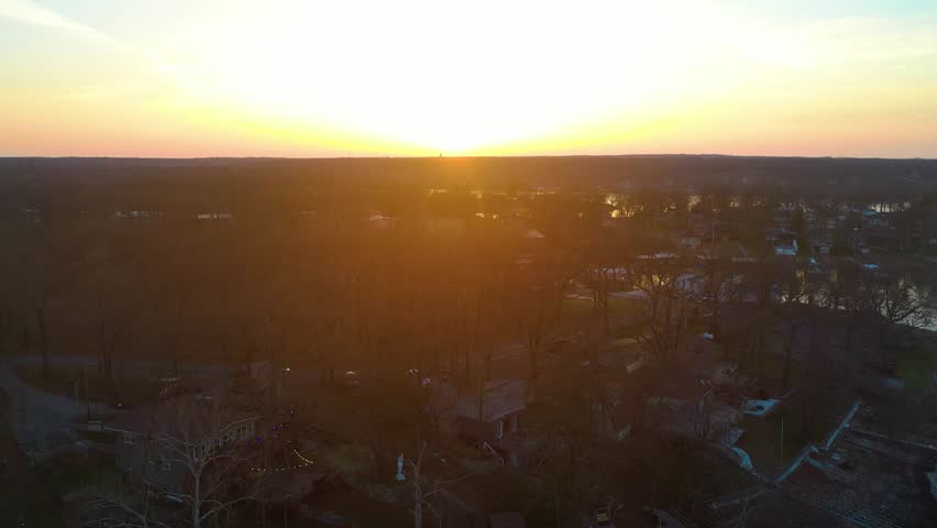 Wide aerial shot near Branson, Missouri, with bright sunset over calm Ozark lake water, forested shoreline, distant homes, soft haze, and golden reflections across the reservoir.