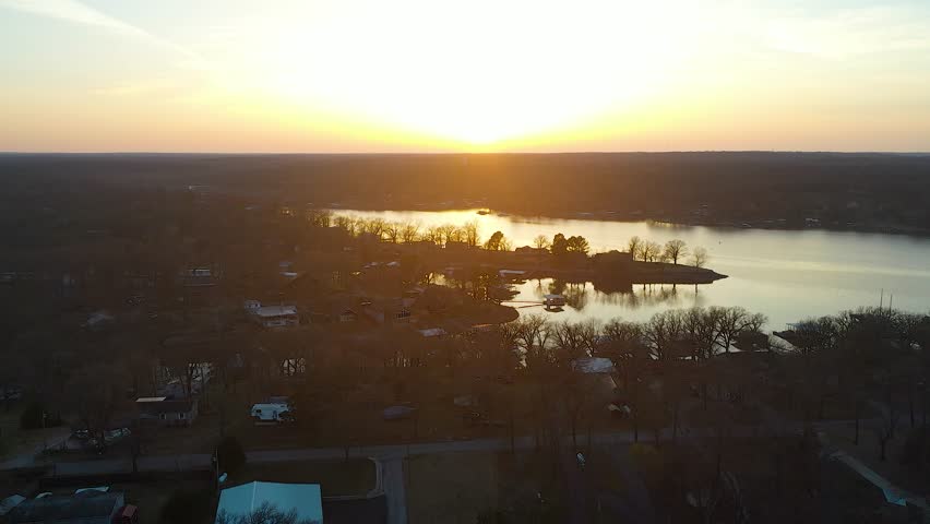 Scenic drone aerial over Beaver Lake, Arkansas at sunset, showing calm rippled water, forested Ozark shoreline, distant lake homes, blue sky, and bright golden sun reflections.
