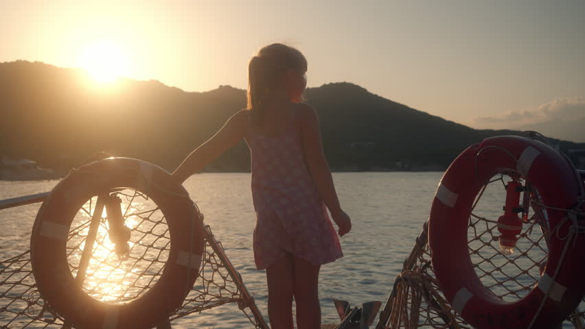 Child watches sunset over water from boat while holding on to lifebuoy near the shore in a quiet location during evening hours