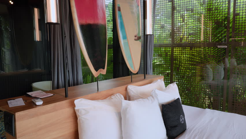 Stylish boutique hotel bedroom featuring comfortable white bedding with fluffy pillows and decorative surfboards mounted on stands behind wooden headboard with lush green tropical foliage visible.
