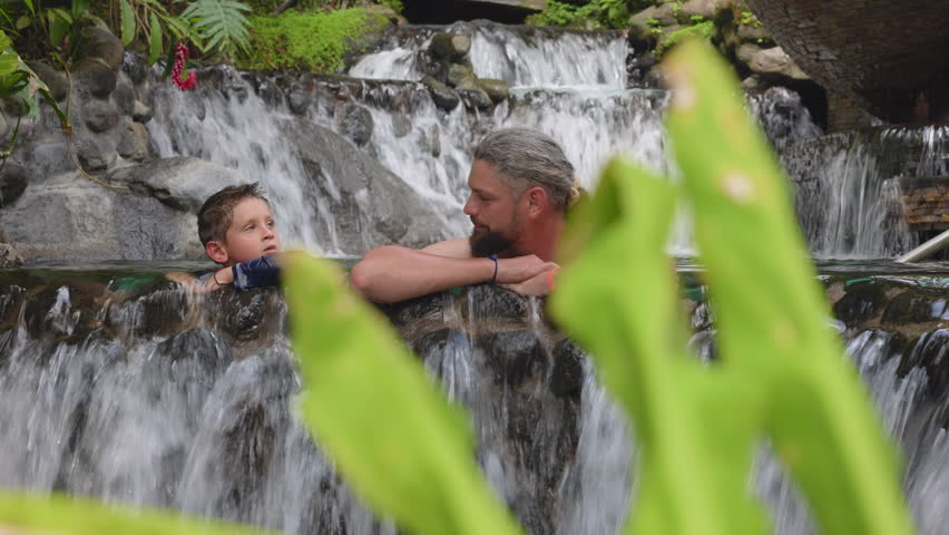 Father and young son share a peaceful moment relaxing together in a natural hot springs pool with tiered waterfalls cascading over volcanic rocks, framed by vibrant green tropical leaves in.