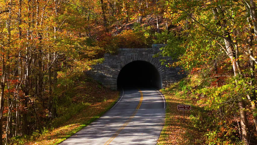Scenic mountain parkway road passing through tunnel beneath rocky hillside with colorful autumn forest. Peaceful seasonal landscape perfect for road trip concepts.