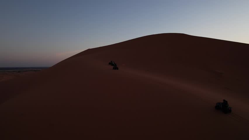 Quad bikes cross the Erg Chebbi dunes near Merzouga, Drâa-Tafilalet, Morocco, beneath towering Sahara ridges illuminated by warm sunset light, drone orbit tracking shot.