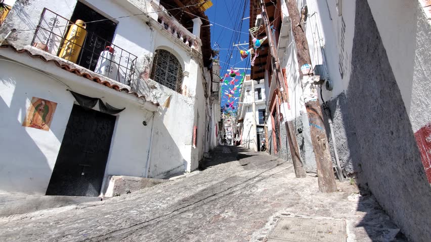 View of steep cobblestone street with white colonial buildings and hanging decorations in Taxco, Guerrero, Mexico.