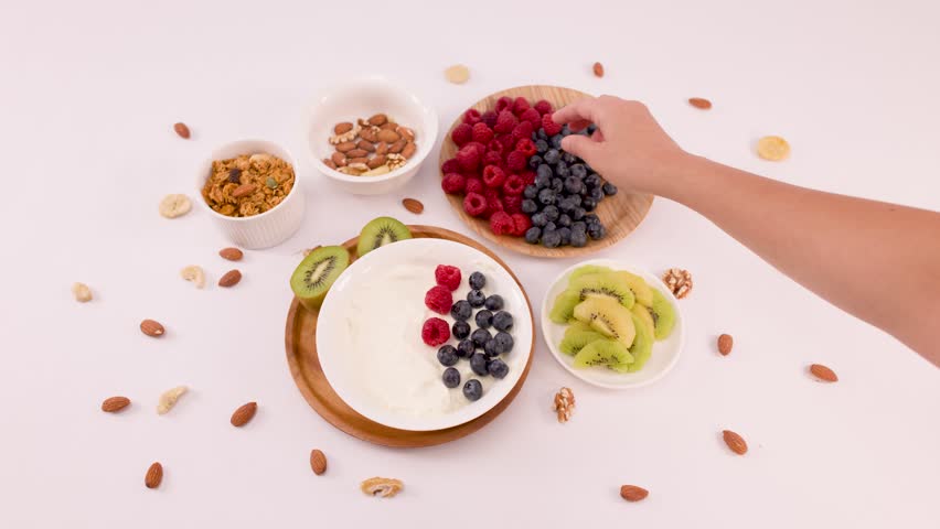 Overhead view of hand adding red raspberries to yogurt bowl with blueberries and kiwi.