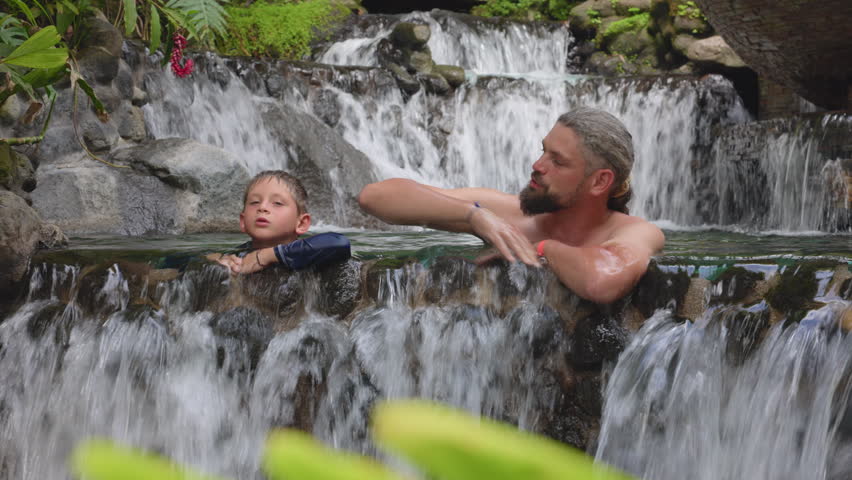 Father and son relax together in natural hot springs pool surrounded by lush tropical vegetation. Warm water cascades over rocks as they share quality time in this serene spa setting.
